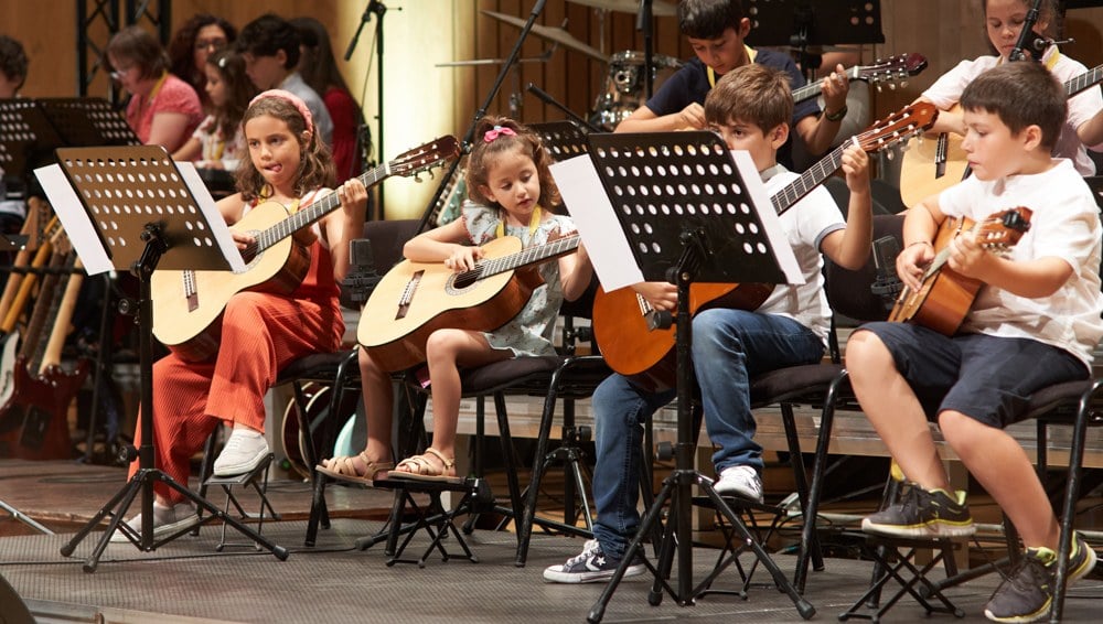 Grupo de crianças em aula de guitarra clássica, concentradas nos atris, numa aula d'Os Músicos